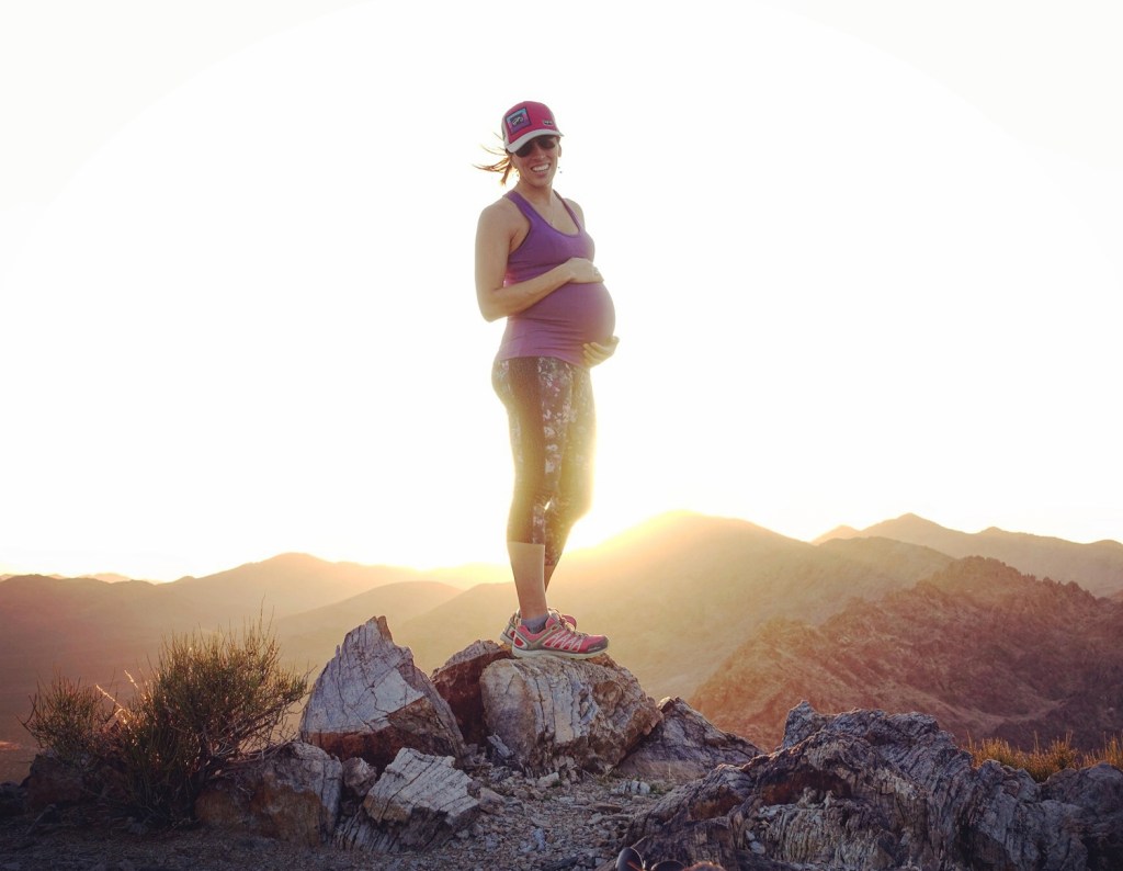 woman stands on mountain top showing her pregnant belly