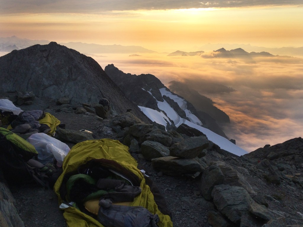 bivy high on a mountain ledge overlooking a sky of clouds and mountain peaks.