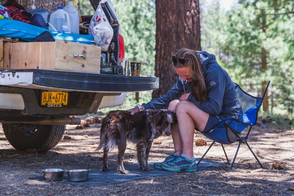 Sarah sits in a camp chair next to her truck's tailgate, brewing coffee and petting her dog.