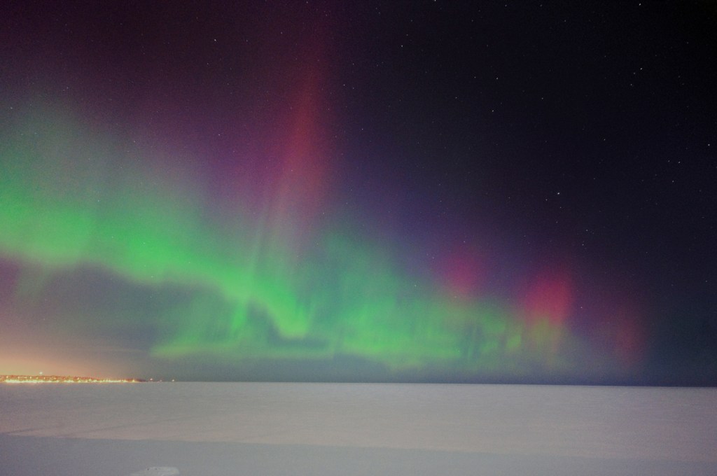 Aurora borealis over a frozen Lake Superior with the lights of Marquette on the shore.