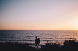 A lone backpacker stands in front of the ocean as the sun is setting.