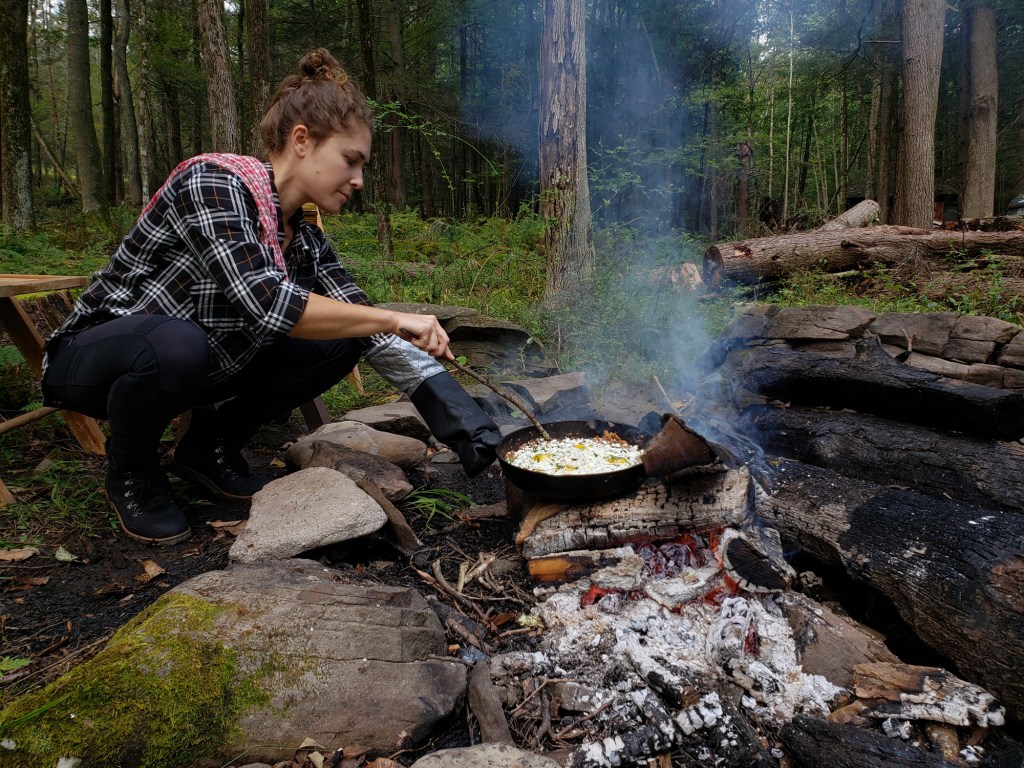 A woman cooking eggs over an open flame in a cast iron pan.