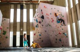 A climber attempts a bouldering problem at an indoor gym, while two other climbers watch and spot.