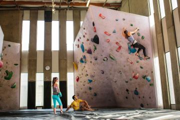 A climber attempts a bouldering problem at an indoor gym, while two other climbers watch and spot.