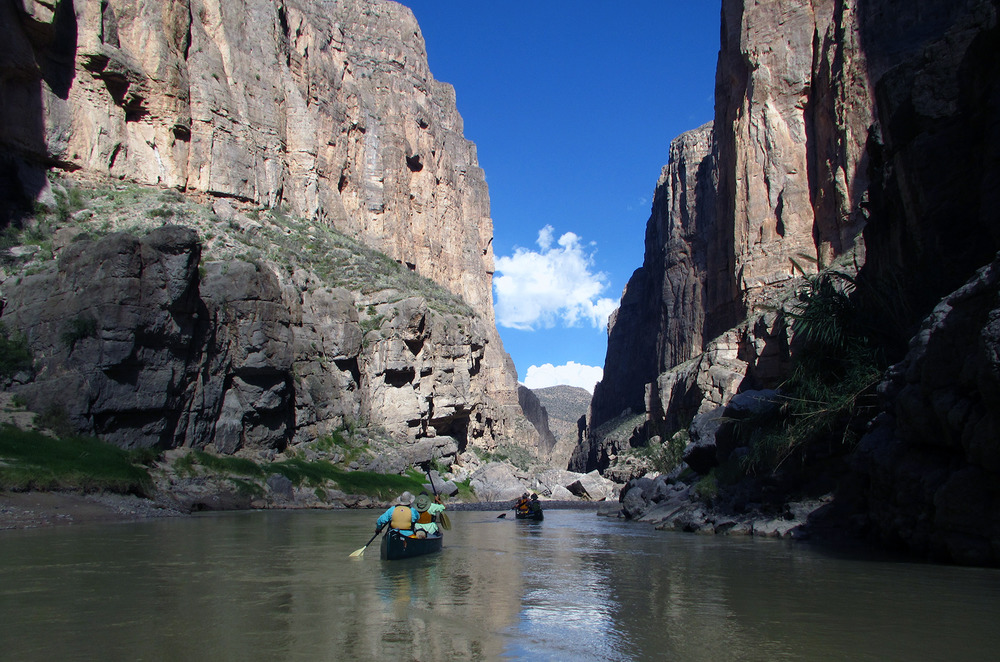 With towering canyon walls overhead, a group of boaters navigate the calm waters of the Rio Grande.