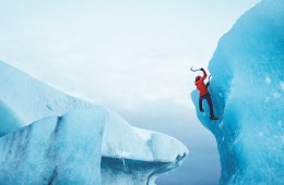 Person in red jacket swings ice axe while climbing a glacier.