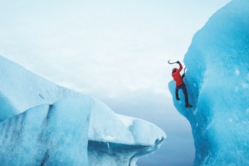 Person in red jacket swings ice axe while climbing a glacier.