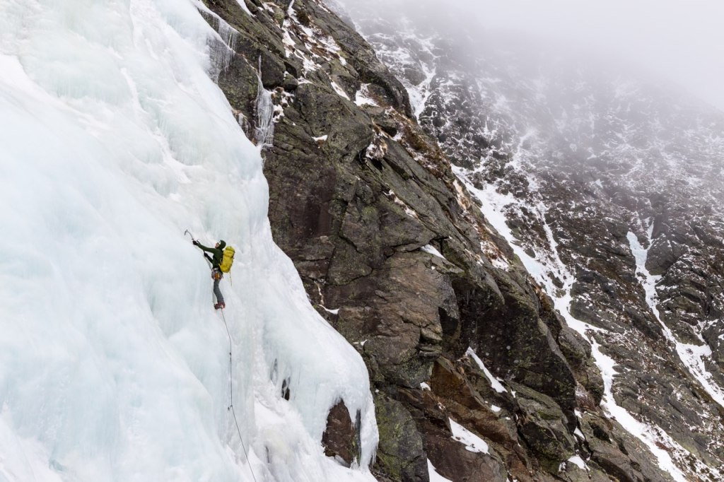 An ice climber makes his way up a frozen waterfall, reaching his ice axe for the next placement.