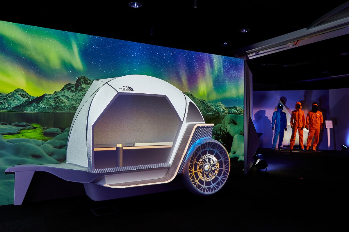 BMW Camper concept stands in front of a mountain scene with the aurora borealis. There are three mannequins in hardshell outerwear in the background.