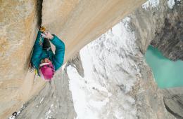 Brette Harrington climbs a deep crack up a rock wall, high above the ground with a turquoise lake below.