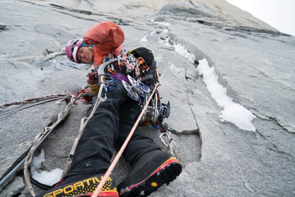 Brette Harrington hangs on a steep rock face, looking through climbing gear strung from her harness. Ice fills the cracks in the rock next to her.