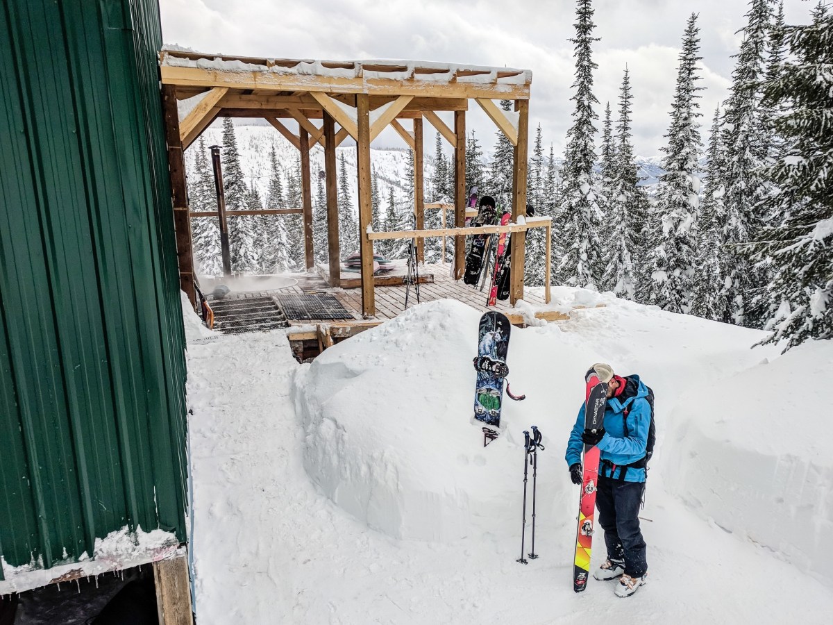 A skier stands with skis outside a hut.