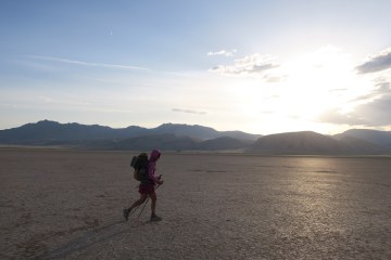 Thru hiker with pack on walks across cracked desert, mountain in the background.