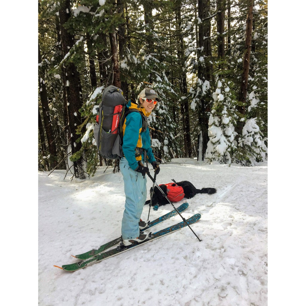 A woman standing on skis in a snowy forest with a heavy pack on her back.