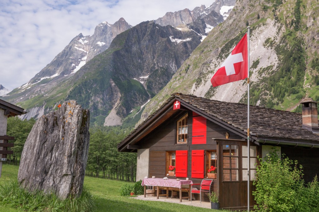 A small building bearing the Swiss flag sits below soaring peaks and rock walls. 