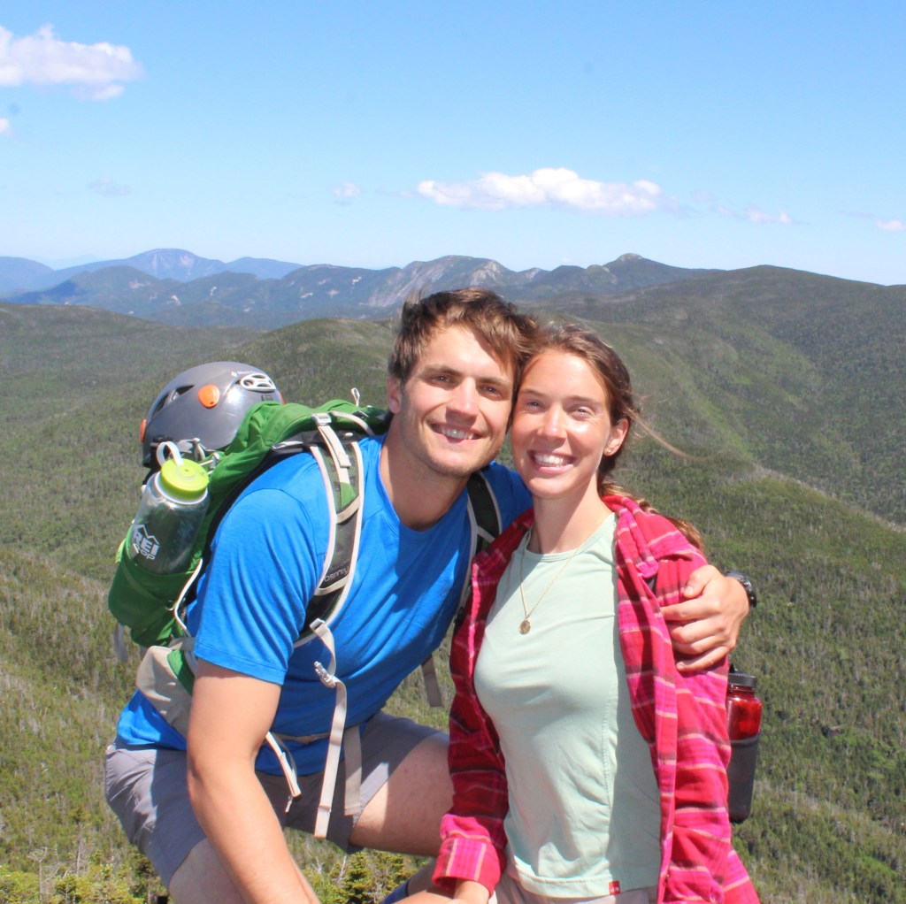 The couple hugs while wearing climbing gear in fronts of a backdrop of green mountains.