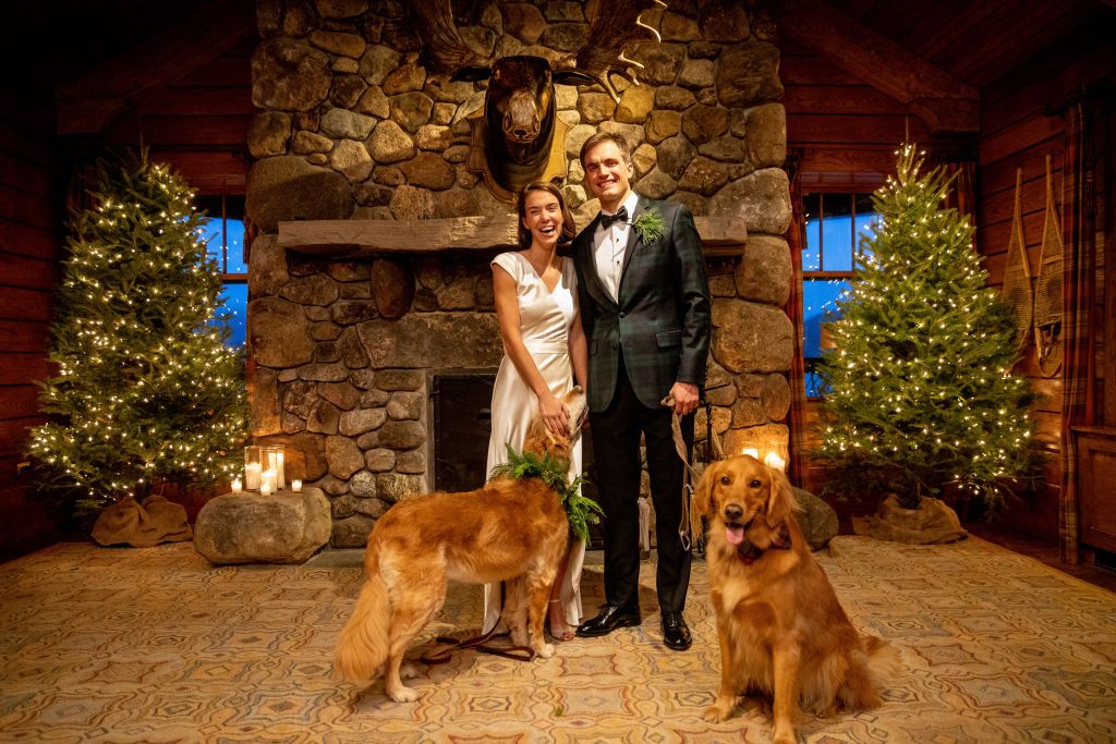 The couple on their wedding day with two golden retrievers in front of a stone fireplace.