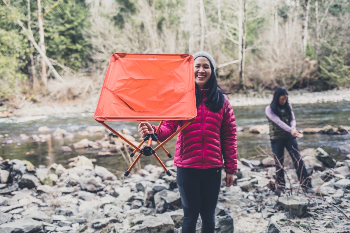 Camper in pink jacket holds orange Flexlite chair in the air.