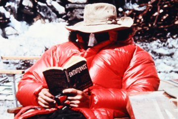 A rock climber on the 1963 American Mount Everest Expedition lounges in a big, red puffy jacket, reading the book "Endurance."