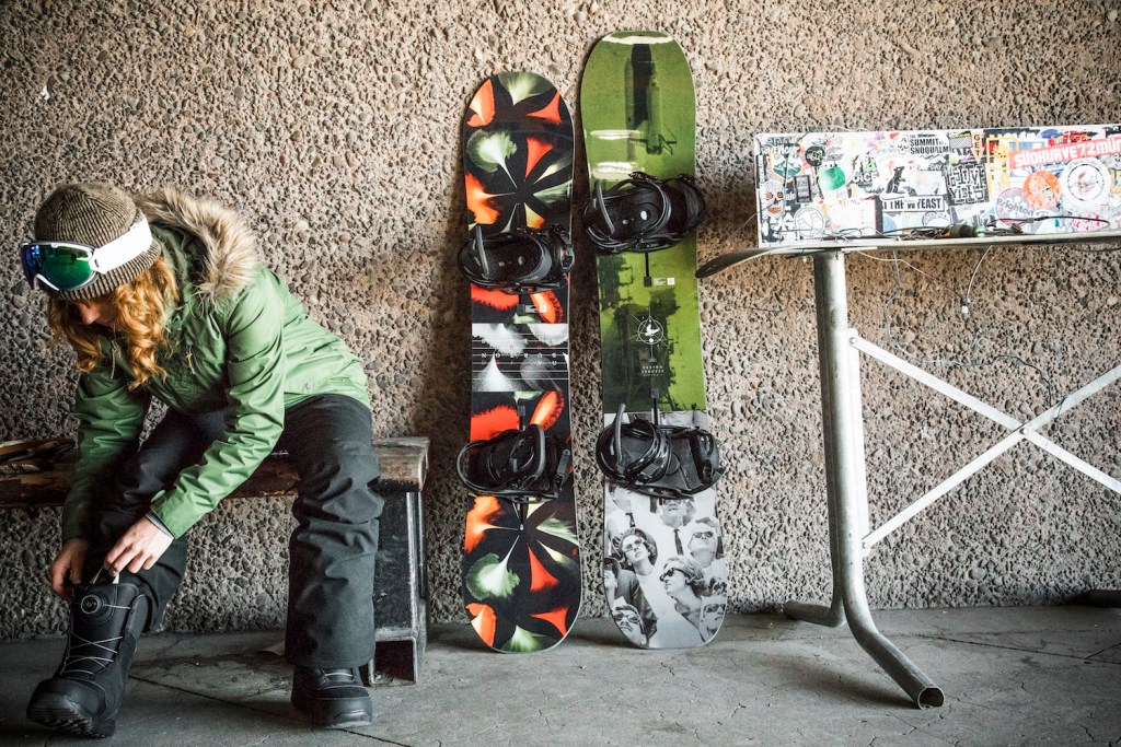 A snowboarder wears bib pants while adjusting boots.