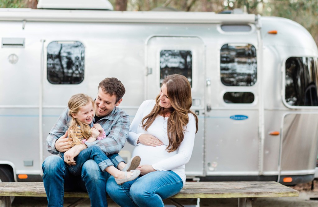 A pregnant mom and dad pose in front of their Airstream with their young child