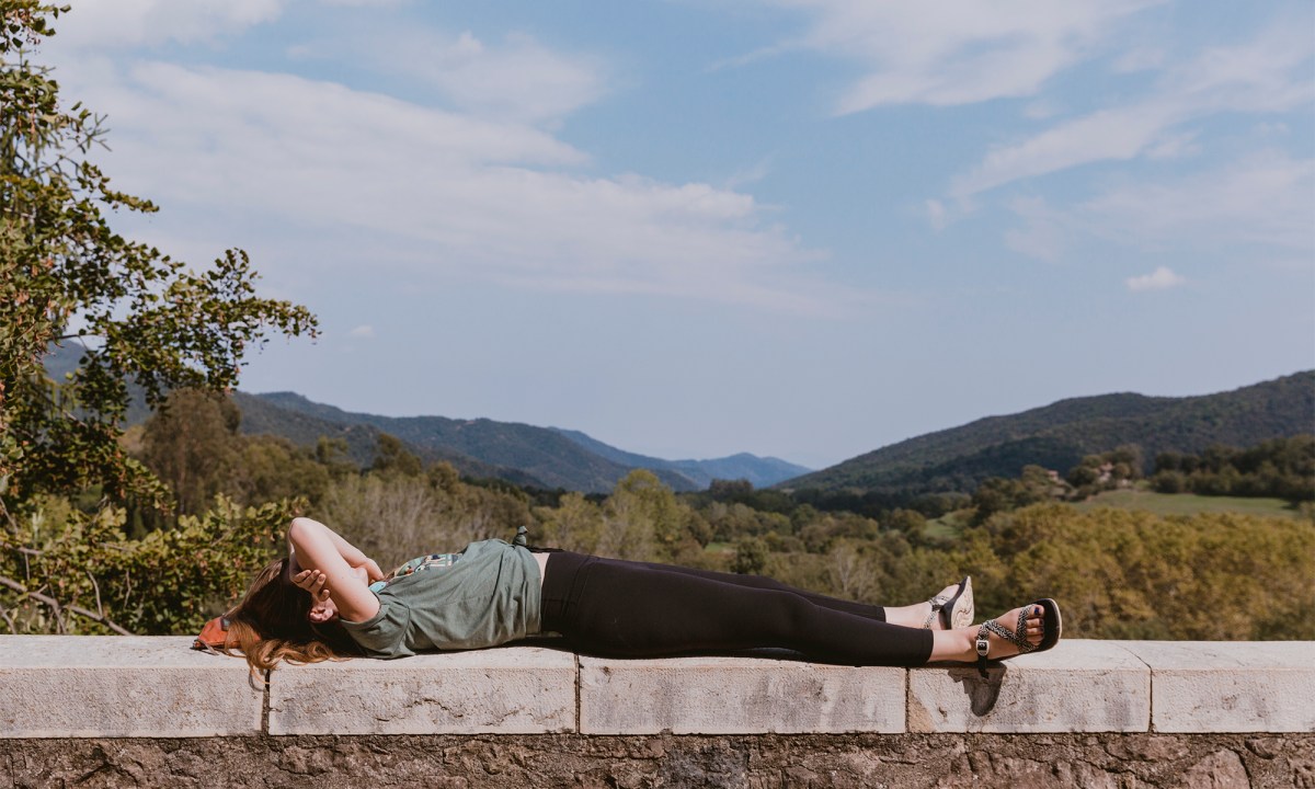 A person lies down on a short wall in front of an open, green vista.