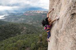 Olivia Hsu reaches dynamically for a hand hold on a steep rock cliff with a green valley stretching out below her.