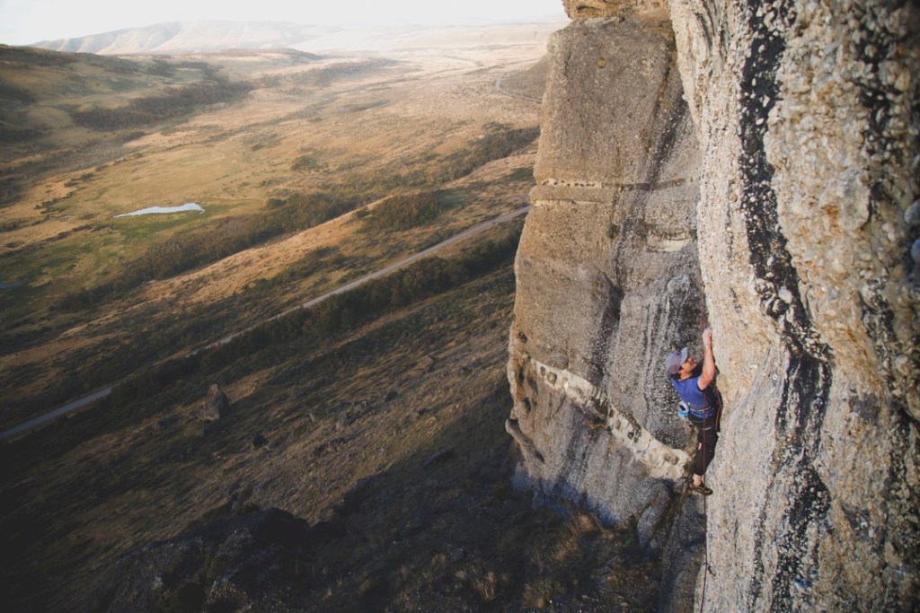 A climber reaches for a hand hold on a cliff face that's bumpy with stone cobbles. A broad valley edged in fog stretches into the background.