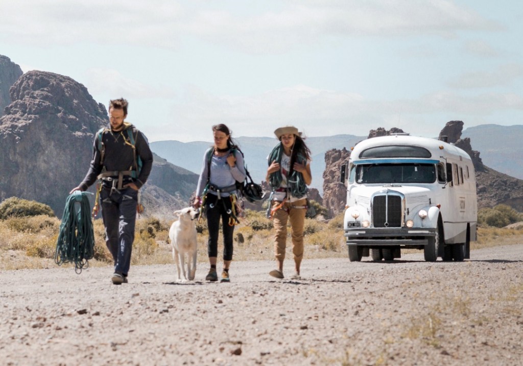 Three people carrying ropes and climbing gear walk along a dirt road on a sunny day with a bus and a mountain in the background.