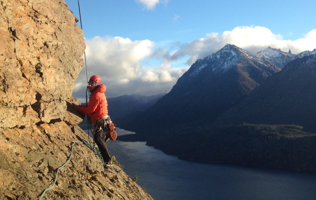 A climber in a red jacket stands on a rock ledge in early morning light with a lake and mountain in the background.