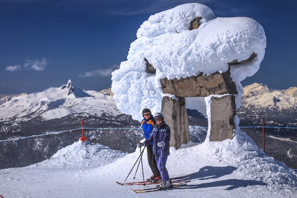 Skiers pose on the Whistler Summit with an inukshuk statue and the Black Tusk looming over the horizon.