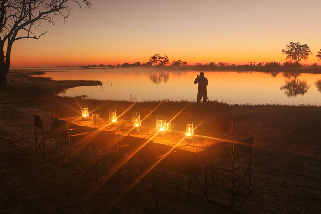 An outdoor dinner table shines from candlelight as the sun sets over a nearby pond.