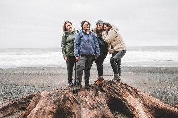 Four hikers stand close on top of a stump near the coast, with gray clouds in the distance.