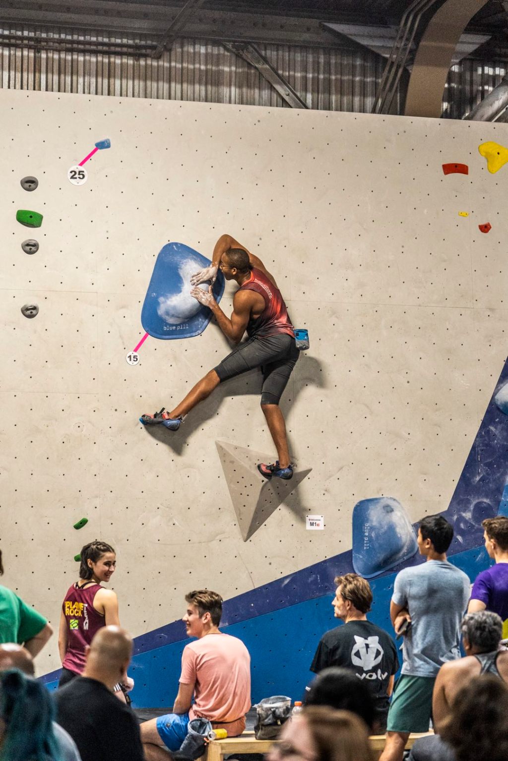 Kai Lightner balances on a pyramid-shaped foothold in a climbing gym, grasping onto a rounded diagonal hold with his hands.