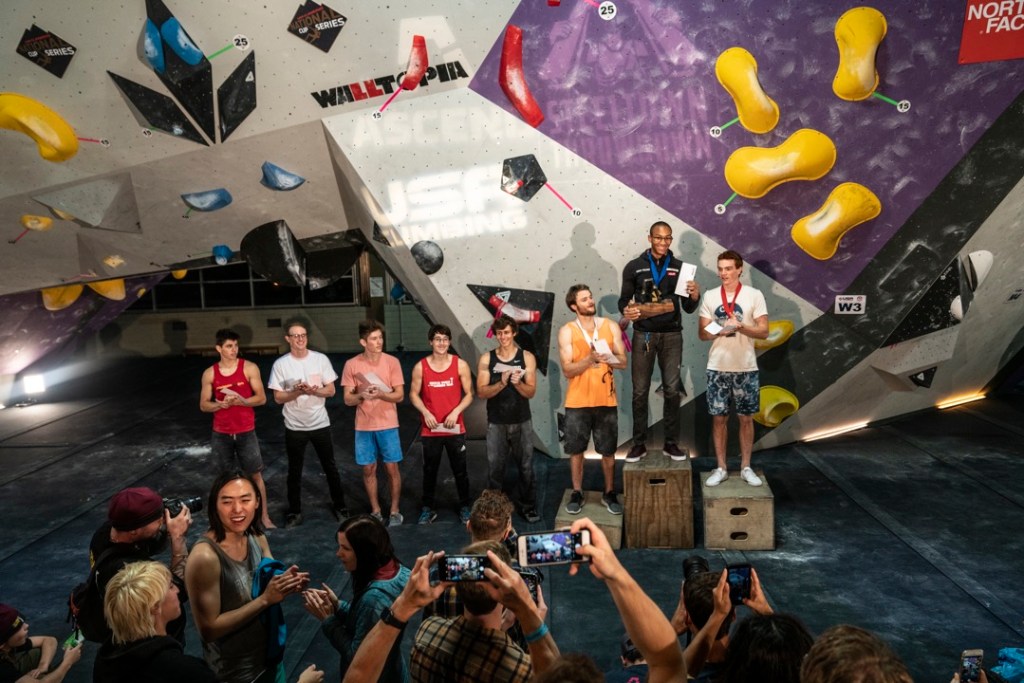 Kai Lightner stand on a podium with a medal around his neck and trophy in his hands at a competittion inside a climbing gym. Two other climbers stand on the lower podium steps and five more line up to the left while an audience applauds and takes photos.