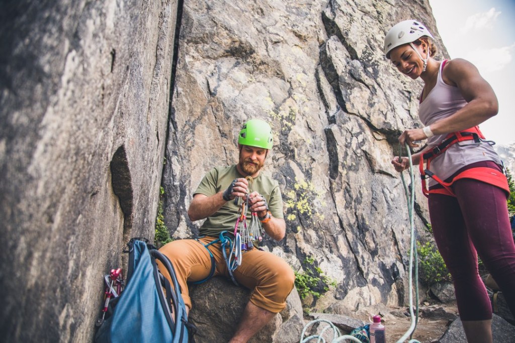 One climber in a helmet sits down to sort through carabiners and climbing nuts while another rock climber stands to sort out a climbing rope.