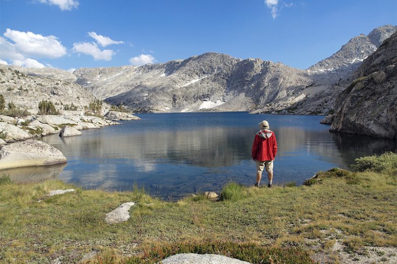 A lone hiker stands in front of an alpine lake deep in the Sierra backcountry.