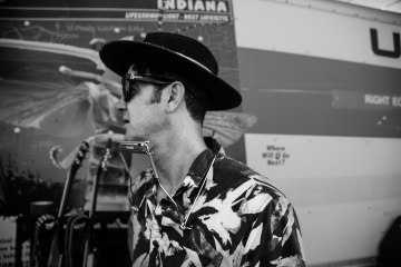 A black and white photo of G-Love facing away from the camera with a harmonica around his neck.