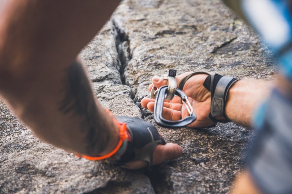 A rock climber wearing protective gloves holds onto a crack in a rock with one hand while inserting a cam for protection in the crack with the other hand.