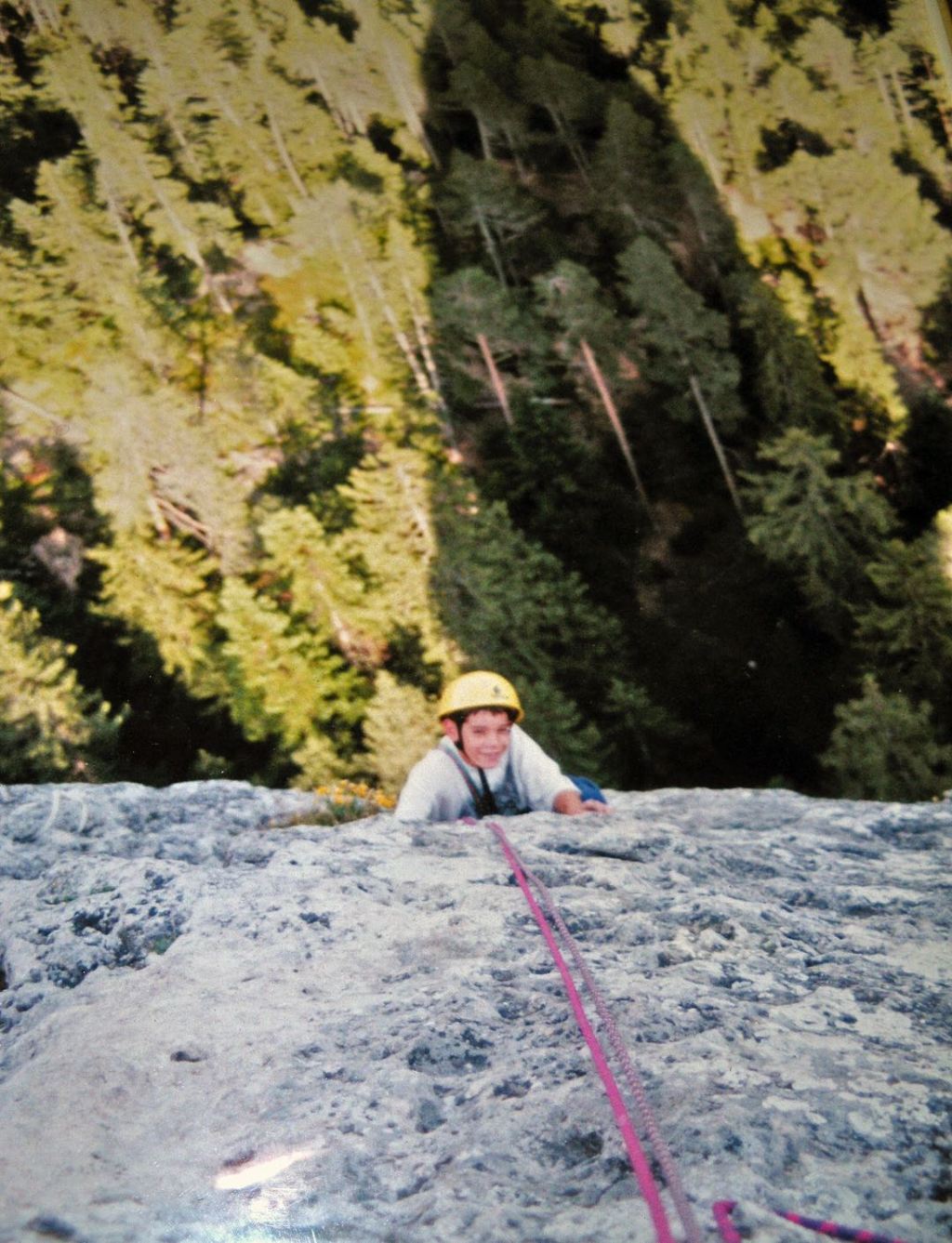 Alex Honnold, as a small boy in a yellow helmet, climbs a steep rock face with evergreen trees stretchingo out far below him.