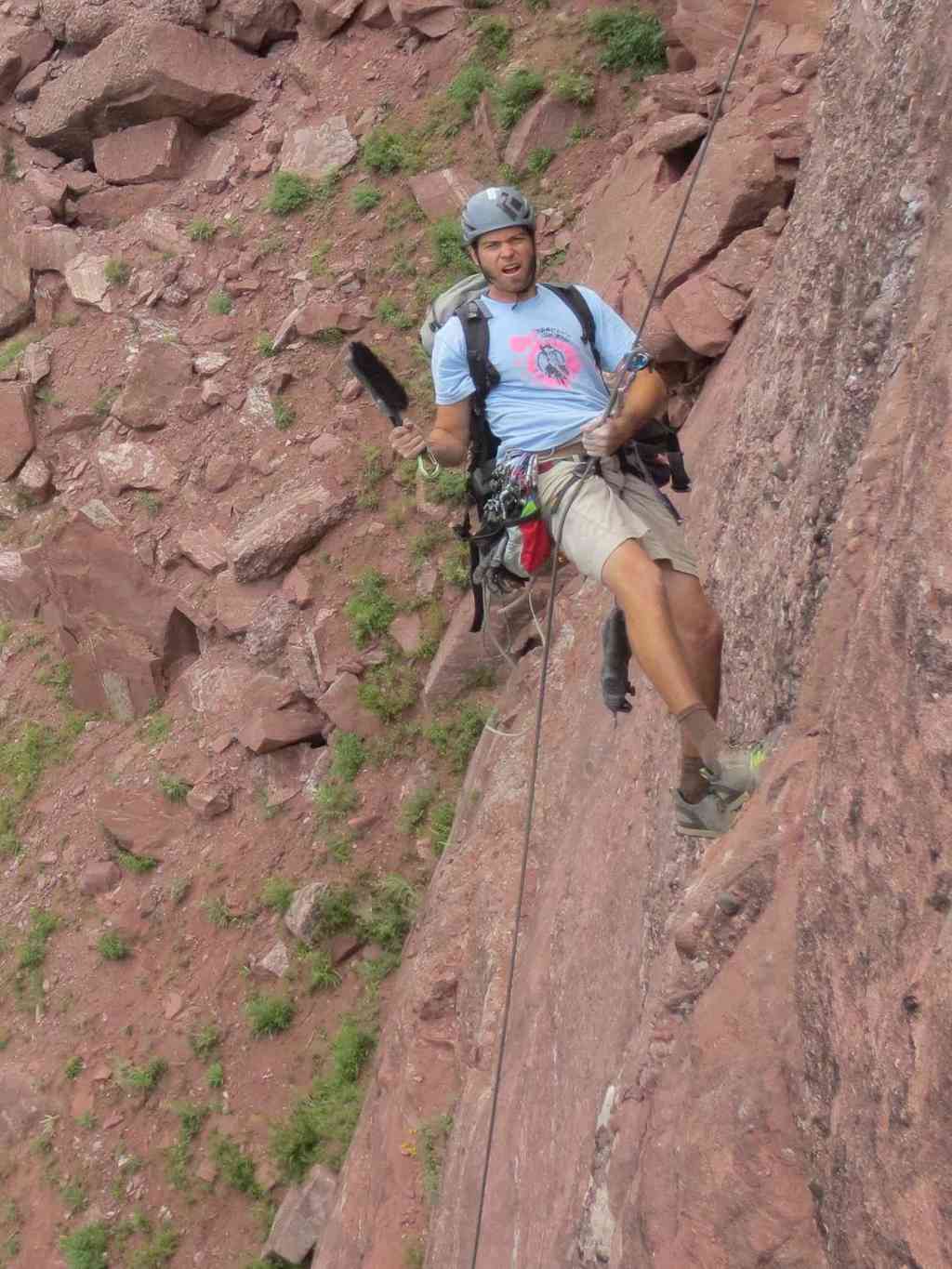 The author, loaded down with a backpack and several items clipped to his harness, waves a small cleaning brush while rappeling down a new climbing route he is developing.