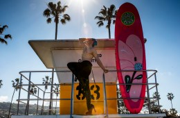 Pattie Gonia on a surf tower in high heeled boots with a pink surfboard behind her.