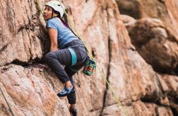 A Rock climber in a white helmet leans in close to a rock wall, stepping one foot high onto a ledge.