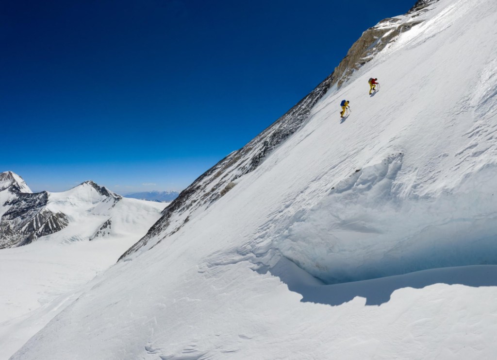 Two climbers, unroped, ascend a steep snow field with a bright blue sky in the background.