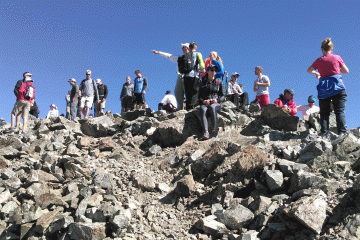 Hikers stand along the top of a ridgeline, looking and pointing every which way under a bright-blue sky.