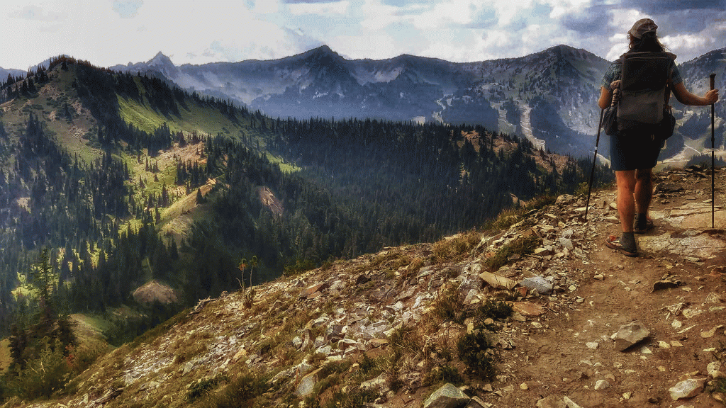 Heather 'Anish' Anderson walks on an exposed trail toward jagged peaks and white, fluffy clouds in the distance