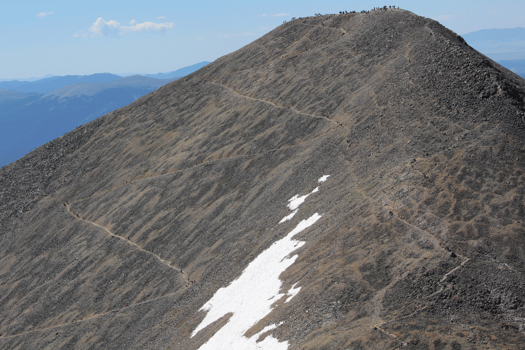 A tall mountain with a bit of snow at the base. The sky is blue and hikers are pictured (they look very small) at the summit.