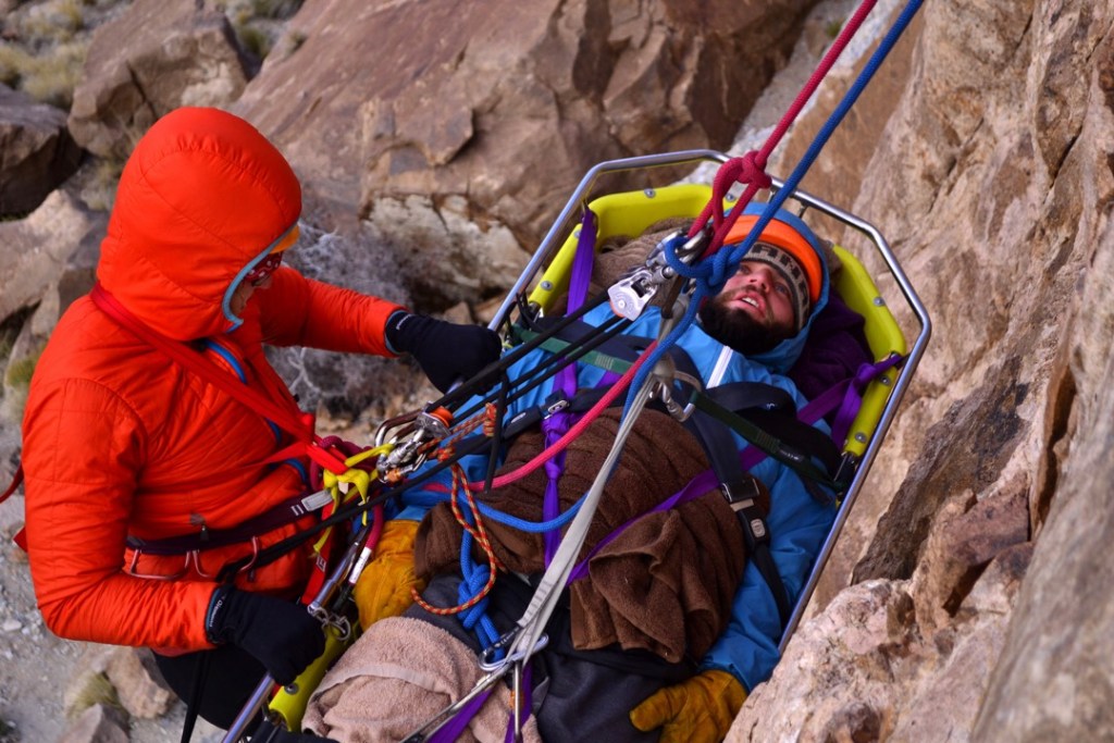 A rock climber is lowered on a stretcher from a steep rock face by another climber.