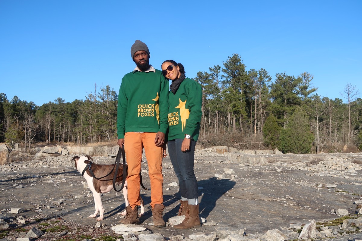 Ayesha McGowan and her husband pose in a rocky field in front of trees with green sweatshirts and their dog. 