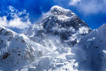 Mount Everest rises high against a blue sky with small, whispy white clouds. Chunks of ice scatter in the foreground.